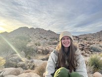 A smiling person sits in front of a mountain covered with scrubby vegetation