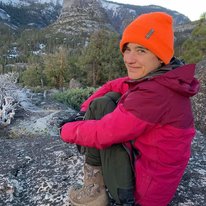 Jess is sitting on top of a mountain with her back to the camera. She is angled slightly facing the camera wearing an orange beanie, pink coat, and brown pants. 