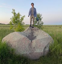Ben is standing on a large boulder in the middle of tall grass. He is wearing a blue and white plaid shirt over a grey t-shirt and khaki pants. 