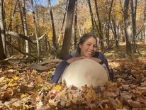 Emily is laying face down in a forest with a lot of leaves on the forest floor. She is facing the camera, smiling with her arms placed on top of a very large puffball mushroom. 
