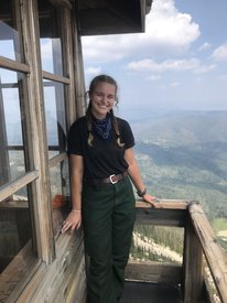 Kylee is standing on a fire lookout platform wearing a black t-shirt and green nomex pants. She is smiling and facing the camera.