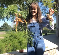 Maria is smiling in her denim overalls while holding a bunch of carrots in her right hand and some onions in her left.