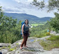 Sarah is standing on a large rock structure with trees and a mountain in the background. She is wearing a blue tank top, brown hiking pants, and a blue hiking pack. 
