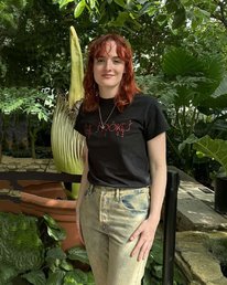 V is standing in front of a Corpse Flower with green leaves from a tree in the background. She is wearing a black t-shirt with acid-washed jeans and facing the camera. Her red hair falls about shoulder-length. 